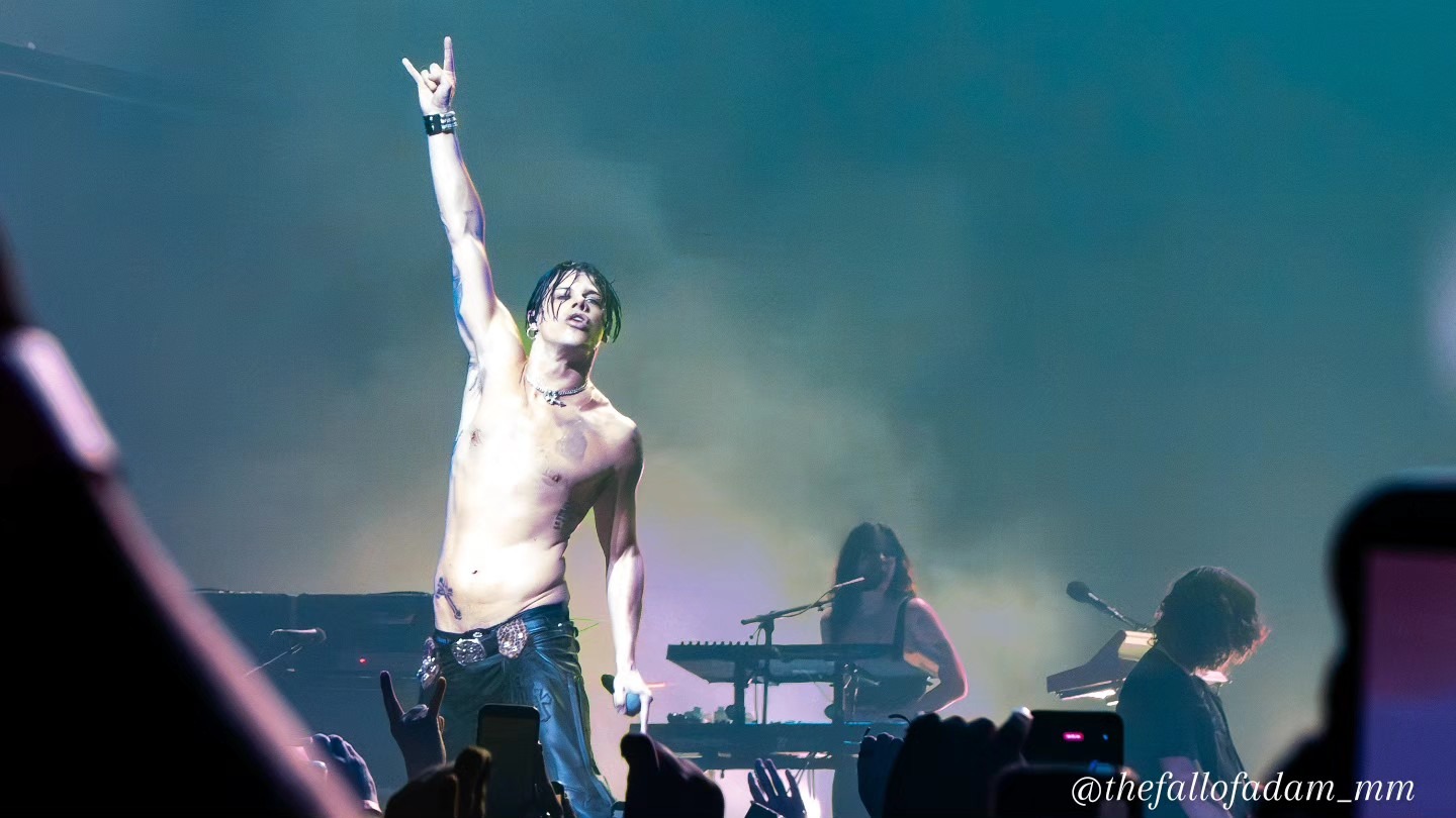 Yungblud raising one arm at the microphone beneath teal light.