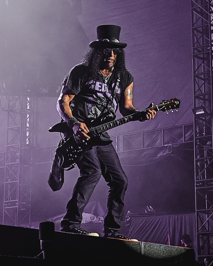 Slash playing guitar in his top hat under purple lighting.